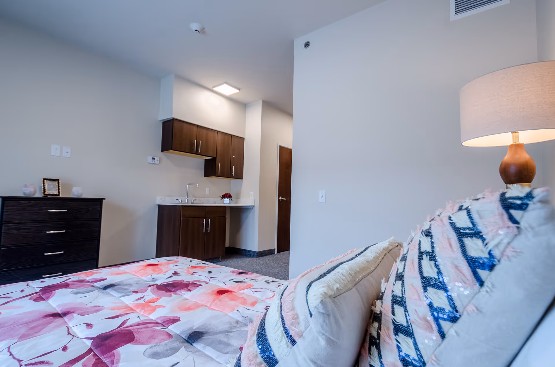 A bedroom in a senior living facility with a bed covered in a floral patterned comforter and decorative pillows. There is a wooden dresser with small decorative items on top, and a kitchenette area with dark wood cabinets and a sink. A lamp with a beige shade is visible on the right side.