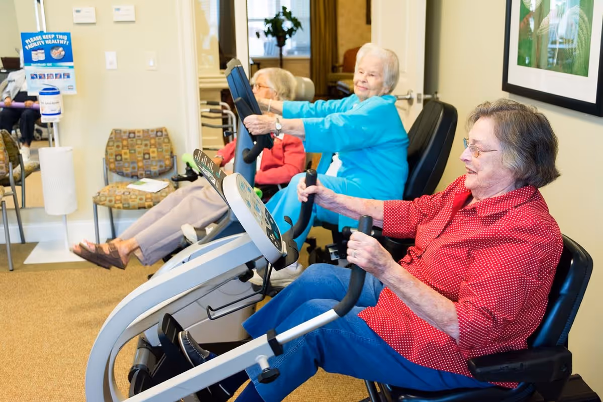 Three elderly women exercising on stationary recumbent bikes in a well-lit room with beige walls and carpet. One woman is wearing a red polka dot shirt, another a turquoise top, and the third a pink jacket. There are chairs and a hand sanitizer station visible in the background.