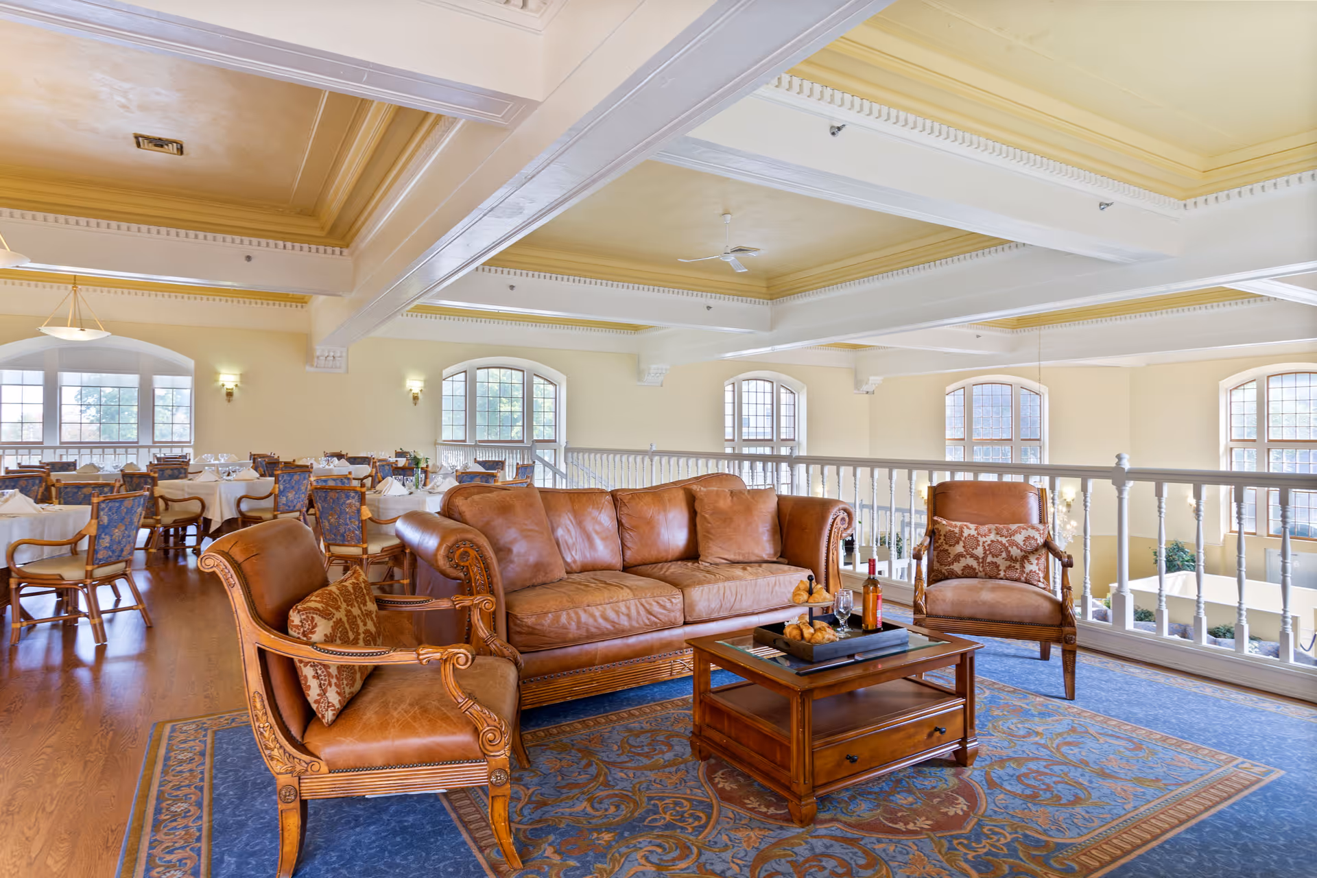 A spacious and elegant common area with a brown leather sofa and two matching armchairs arranged around a wooden coffee table on a patterned blue and brown rug. Behind the seating area, there are several dining tables with chairs set up near large arched windows letting in natural light. The room features a high ceiling with decorative beams and soft yellow walls.