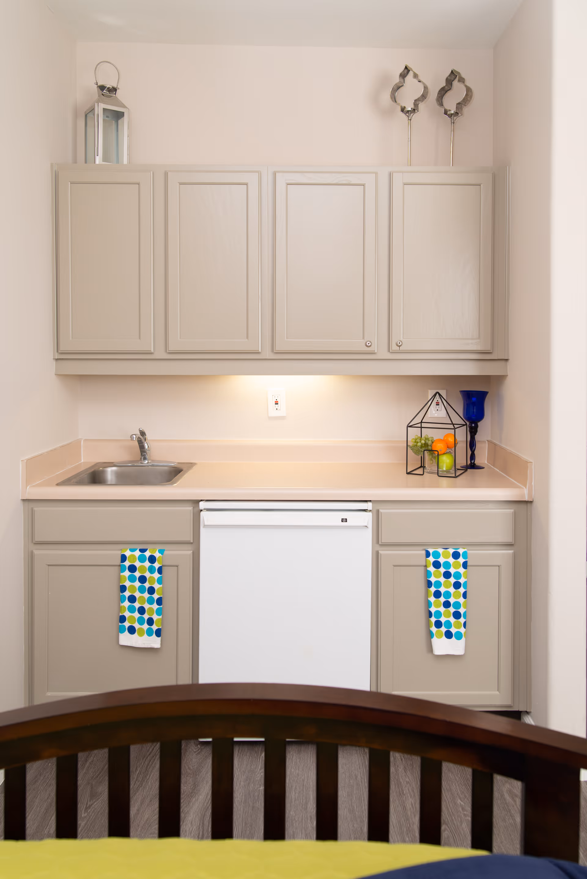 Compact kitchenette with beige cabinetry, a small sink, white under-counter dishwasher, and decorative items on the counter.