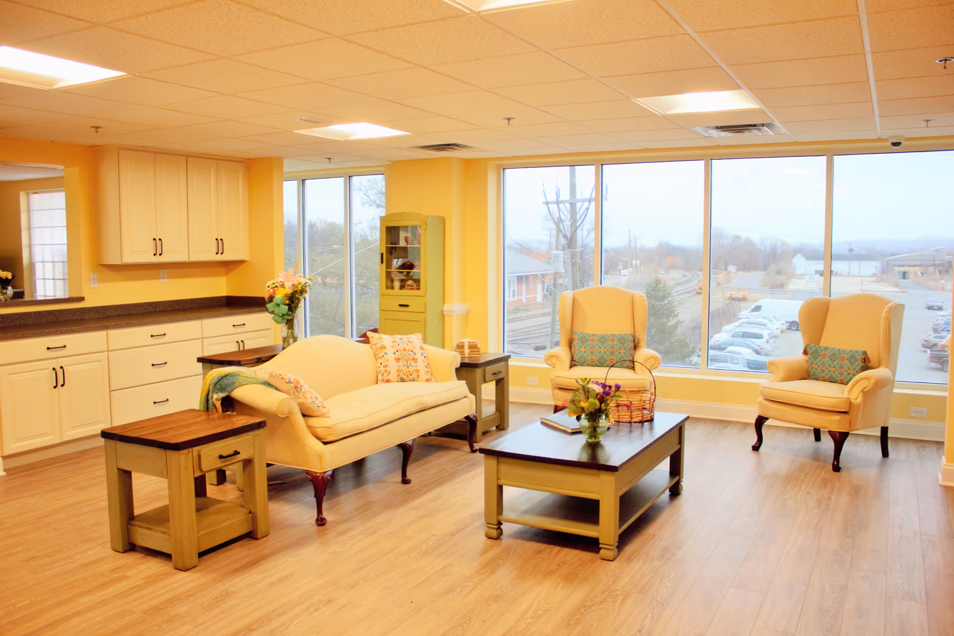 A bright and cozy living room area with large windows showing an outside view of a parking lot and railway tracks. The room features a cream-colored sofa with floral cushions, two matching armchairs with patterned pillows, a wooden coffee table with a flower vase and basket, and side tables. The walls are painted yellow, and there are cabinets and a countertop along one wall.
