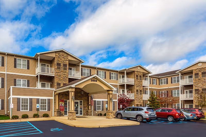 Exterior front view of a three-story retirement community building with beige siding and stone accents. The entrance features a covered portico with the number 3959 above it. Several cars are parked in the parking lot in front of the building, and the sky is partly cloudy with blue patches.