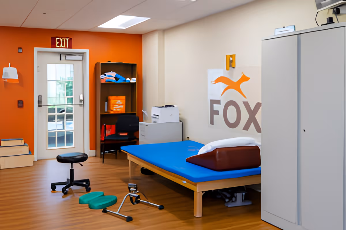A therapy or treatment room with a blue padded treatment table, a black rolling stool, a small exercise pedal machine on the floor, a chair, a wooden shelf with folded towels and supplies, a white cabinet, and an exit door with a window. The walls are painted orange and beige, and there is a large FOX logo on the beige wall.