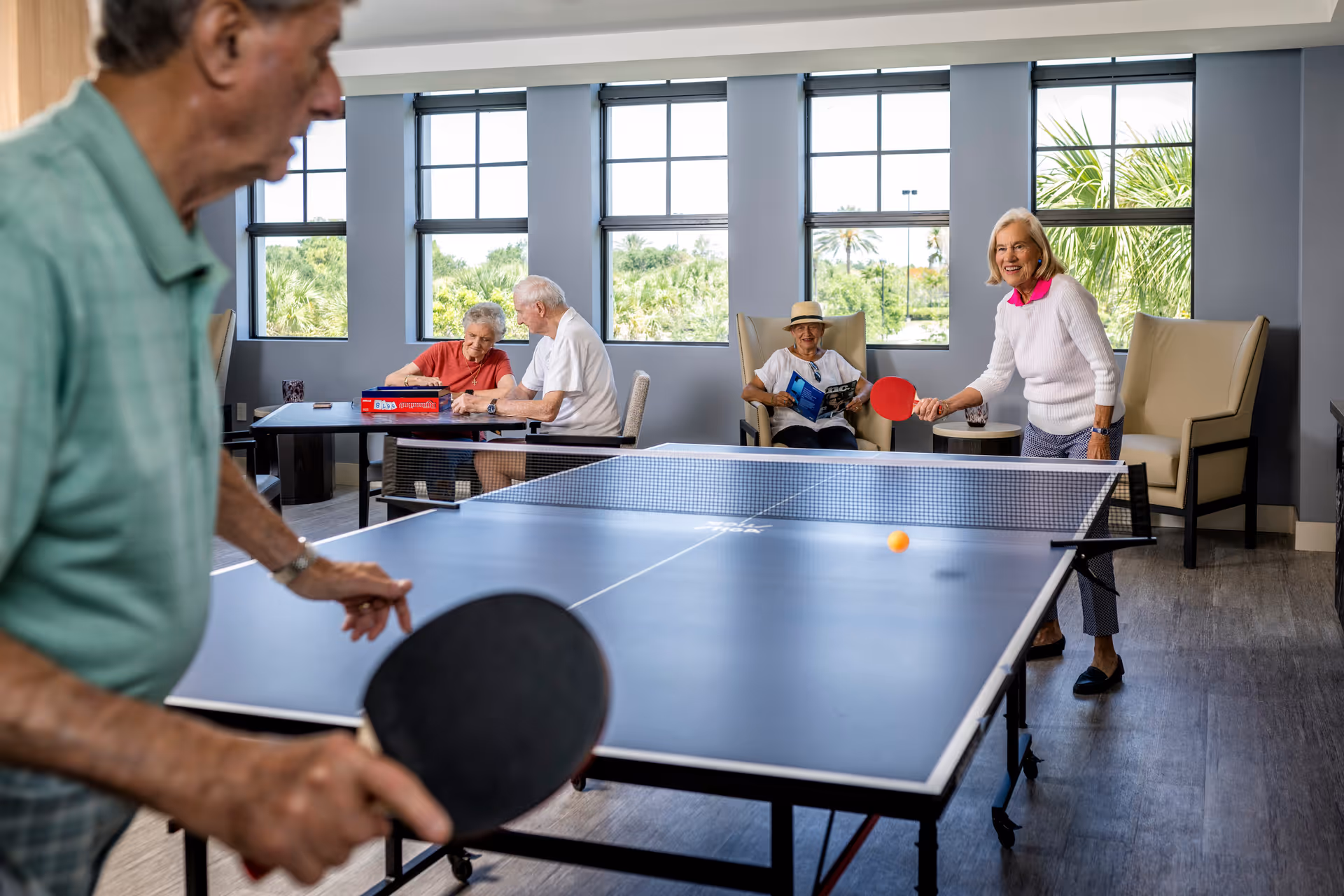 Two elderly people playing ping pong in a bright room with large windows showing greenery outside. In the background, two elderly people are sitting at a table playing a board game, and another elderly person is sitting in a chair reading a magazine.