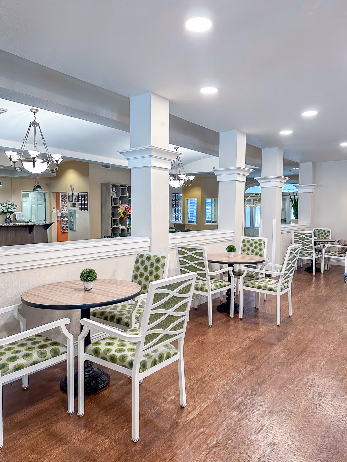 Interior view of a senior living facility dining area with round wooden tables and white chairs featuring green patterned cushions. The space has wooden flooring, white pillars, and ceiling lights, with a chandelier visible in the background.