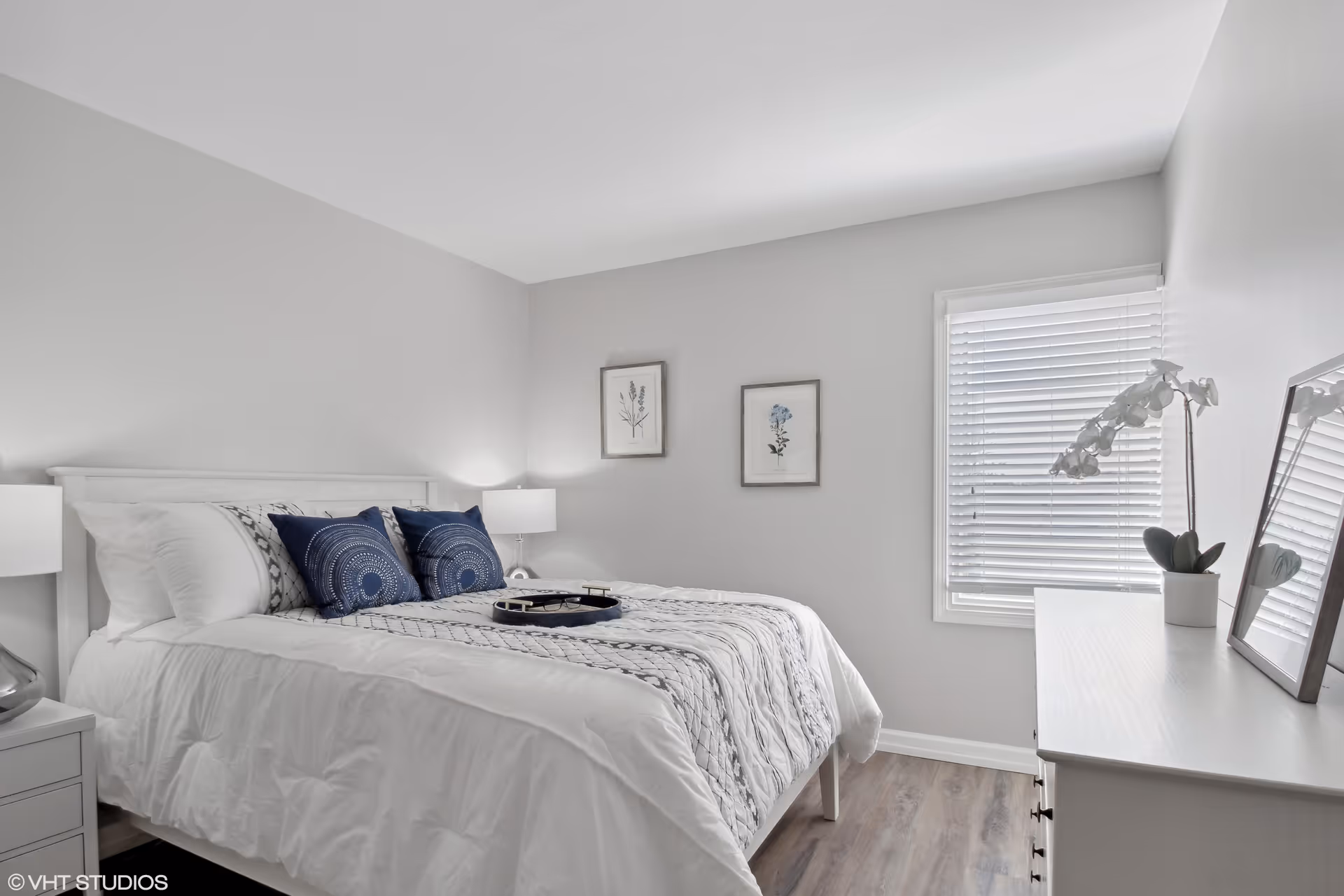 A bright and clean bedroom with a white bed featuring white and blue pillows and a patterned white and gray bedspread. There are two framed botanical prints on the wall above the bed, a window with white blinds, a white dresser with a mirror and a white orchid plant, and two bedside tables with lamps.