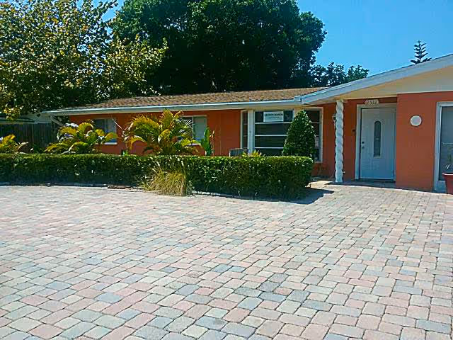 Front exterior view of a single-story building with an orange facade, a white door, and windows. There are neatly trimmed bushes and palm plants in front of the building, and a paved stone driveway or walkway in the foreground. Trees are visible in the background under a clear blue sky.