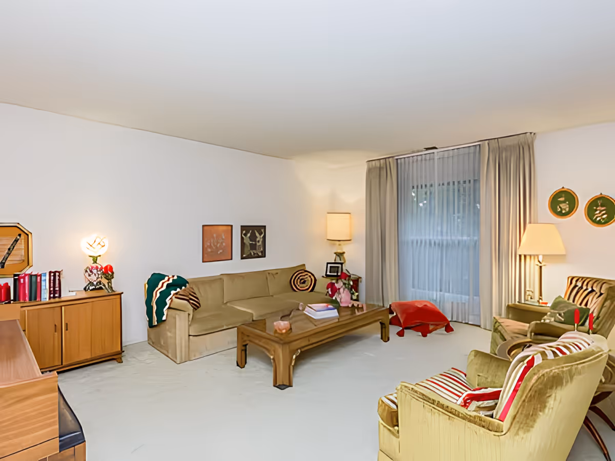 A cozy living room with beige walls and carpet. The room features a long beige sofa with a colorful blanket draped over one arm, two green armchairs with striped cushions, a wooden coffee table with books and decorative items, and two lamps providing warm lighting. There is a large window with sheer white curtains and beige drapes, and various framed artworks and decorative plates on the walls.