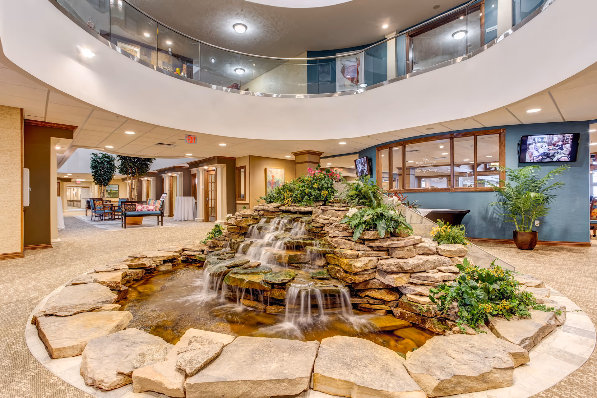 Indoor atrium of a retirement community with a central rock waterfall pond, surrounding plants and seating, and a second-floor curved balcony.