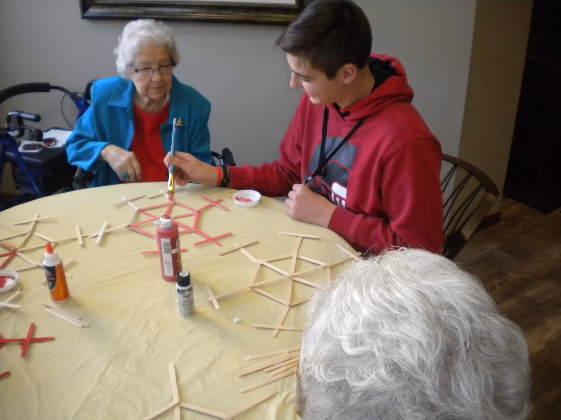 An elderly woman and a young man sitting at a round table covered with a yellow tablecloth, engaging in a craft activity with popsicle sticks and paint. The elderly woman is wearing a blue jacket and red shirt, while the young man is wearing a red hoodie and painting some of the popsicle sticks red. Various craft supplies including glue and paint bottles are on the table.