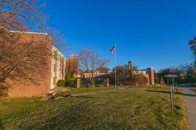 Exterior view of a brick building with a well-maintained lawn, trees, and an American flag on a flagpole under a clear blue sky.