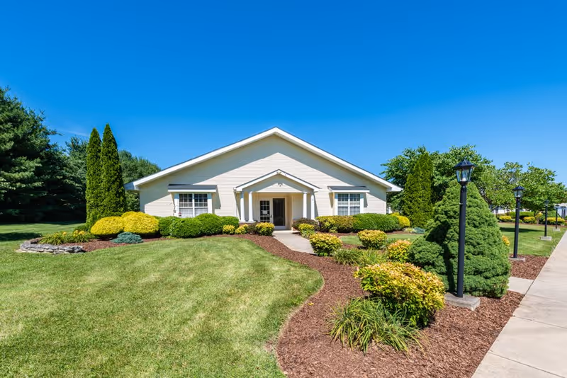 Single-story building with a white exterior surrounded by well-maintained green shrubs and trees under a clear blue sky. A concrete walkway leads to the entrance, flanked by black lamp posts and landscaped garden beds with mulch.