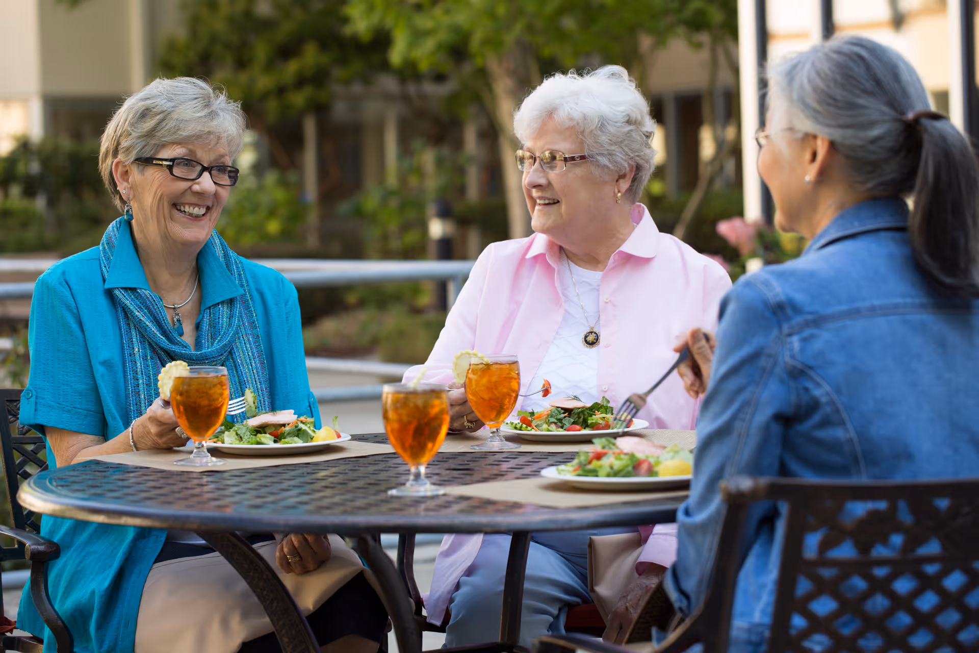 Three elderly women sitting around a metal outdoor table enjoying a meal together. They have plates of salad and glasses of iced tea with lemon. The setting appears to be a garden or patio area with greenery and buildings in the background.