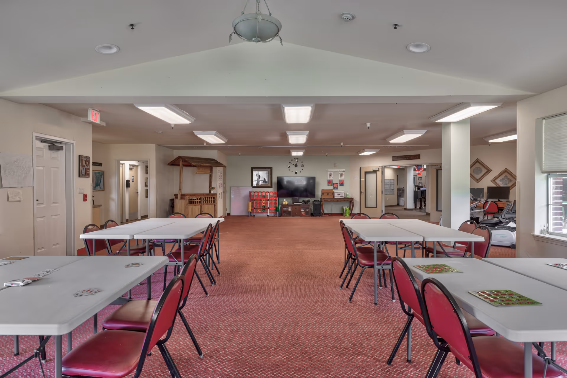 A spacious activity room with several rectangular tables and red chairs arranged in rows on a red carpet. The room has white walls and ceiling with fluorescent lights. At the far end, there is a large flat-screen TV mounted on the wall, a clock above it, and some shelves with games and supplies. There are windows on the right side letting in natural light, and a doorway leading to other rooms on the left and center.