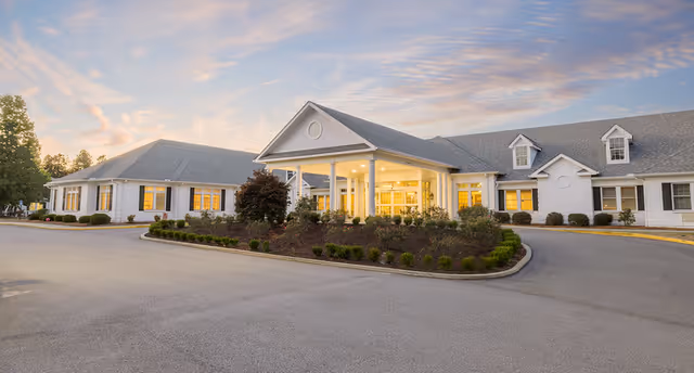 Exterior view of a single-story senior living facility building with a covered entrance, surrounded by landscaped bushes and trees under a partly cloudy sky at sunset.