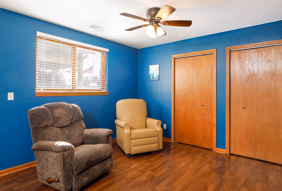 A small sitting room with blue walls, two recliner chairs, a ceiling fan, a window with blinds, and two wooden closet doors.