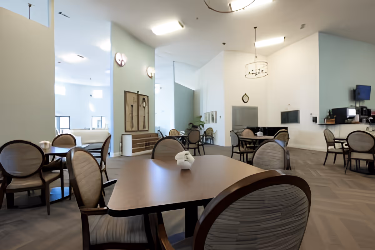 Spacious senior living dining room with multiple round tables and upholstered chairs, chandeliers, and a service counter.