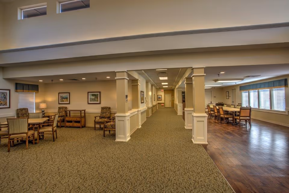 Interior view of a senior living facility common area with a carpeted hallway in the center, seating areas with tables and chairs on the left, and a dining area with a long table and chairs on the right near large windows with blinds.