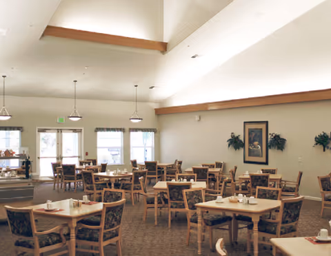 Large dining room with several square tables and upholstered chairs beneath a high vaulted ceiling and pendant lights.