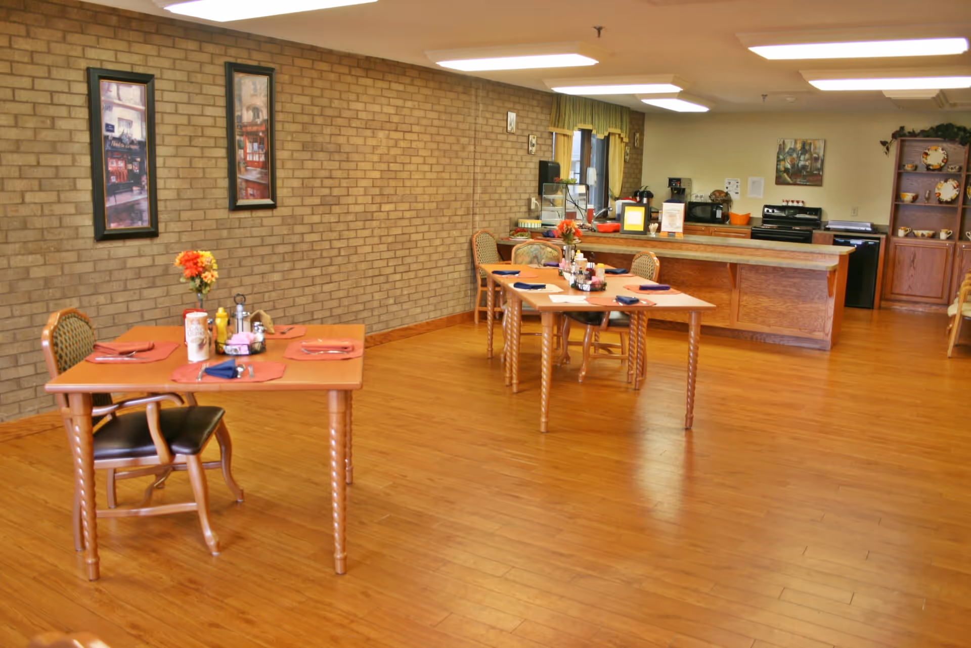 Dining room with wooden tables and chairs set with placemats and condiments and a serving counter/kitchen in the background.