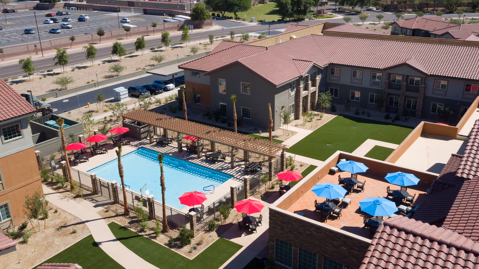 Aerial view of an outdoor pool area at The Enclave at Gilbert Senior Living, featuring a rectangular swimming pool surrounded by a fence, several red umbrellas with seating, a pergola with shaded seating, and an adjacent patio area with blue umbrellas and tables. The surrounding buildings have red-tiled roofs and beige walls, with landscaped pathways and green lawns.