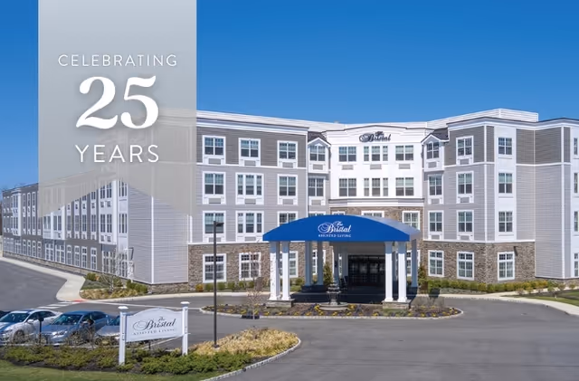 Exterior view of The Bristal Assisted Living at Wayne building under a clear blue sky, featuring a large multi-story structure with white and gray siding, a blue awning over the entrance, and a sign in front. Text overlay on the left side reads 'Celebrating 25 Years'.