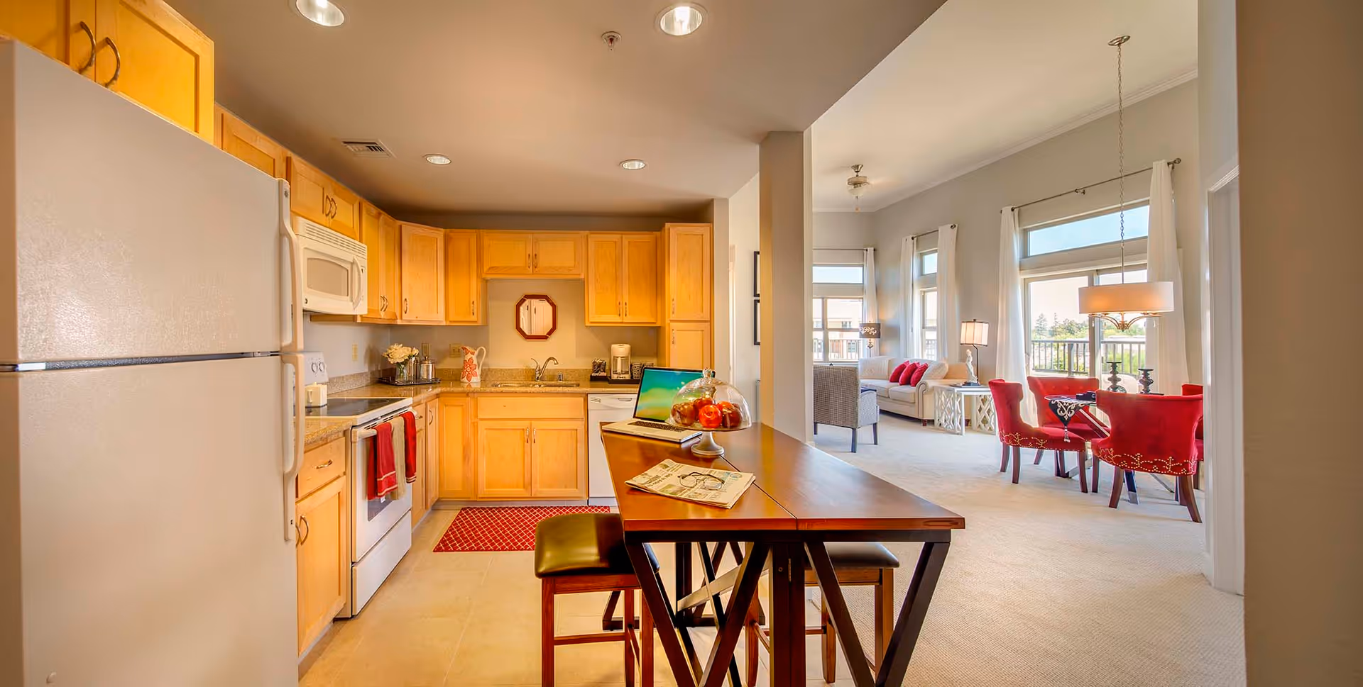 Bright and spacious kitchen with light wood cabinets, white appliances including a refrigerator, stove, microwave, and dishwasher. A wooden table with stools is positioned in the foreground with a laptop, a glass cake stand with pastries, and an open book on it. The kitchen opens into a living area with large windows, white curtains, a white sofa with red pillows, a gray armchair, and a dining area with red upholstered chairs around a dark wooden table under a hanging light fixture.