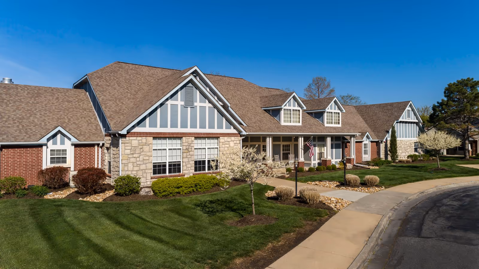 Exterior view of Homestead Assisted Living of Olathe South, showing a large building with a combination of brick and stone facade, multiple gabled roofs, and a well-maintained lawn with small trees and shrubs under a clear blue sky.
