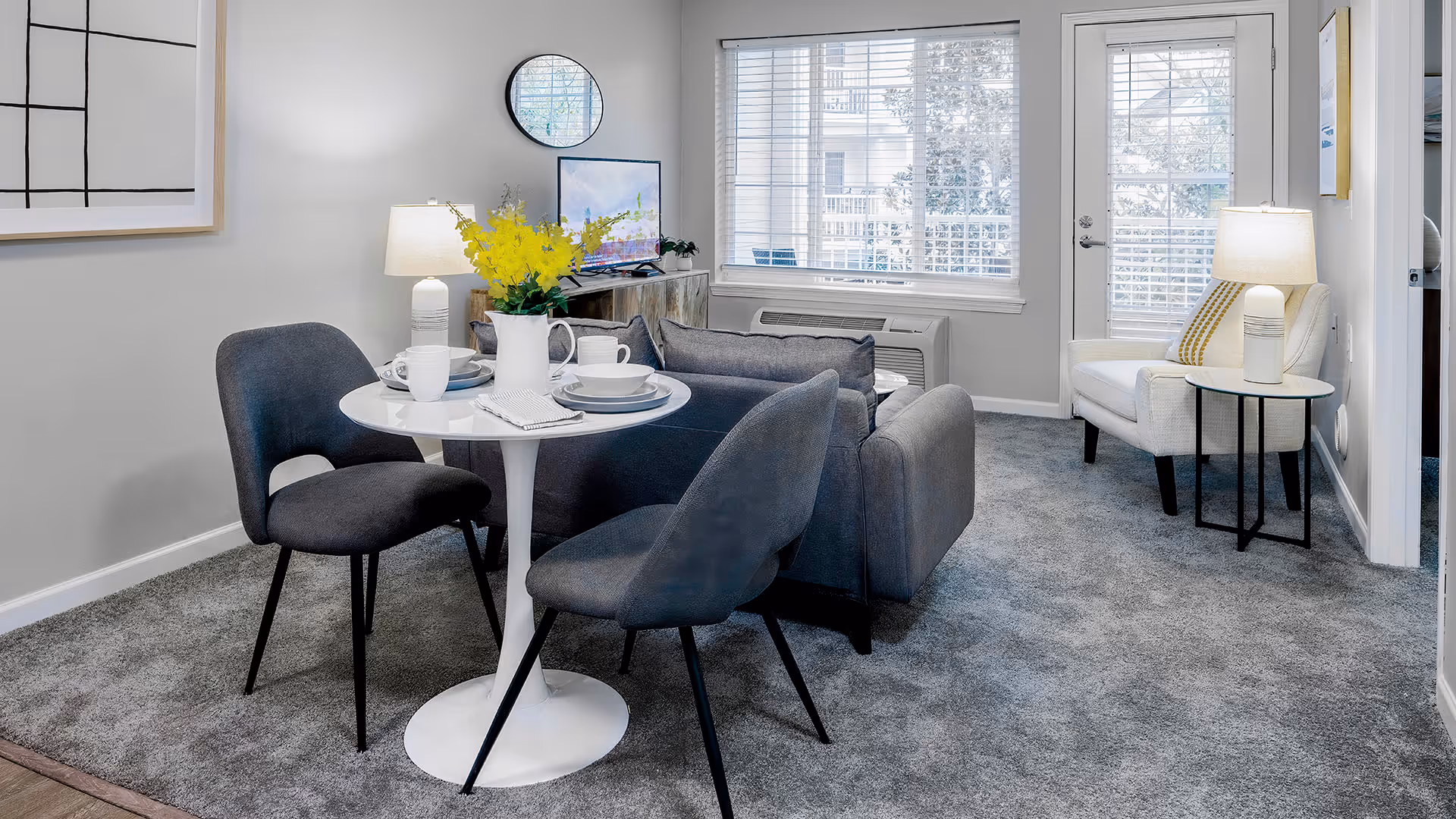 A cozy living room area with a small round white dining table set for two with white dishes and a vase of yellow flowers. Two dark gray upholstered chairs are around the table. Behind the table is a gray sofa facing a TV on a wooden stand. A round mirror and framed artwork hang on the light gray walls. A white armchair with a yellow striped pillow sits next to a small round side table with a lamp. Large windows with white blinds and a glass door let in natural light. The floor is covered with gray carpet.