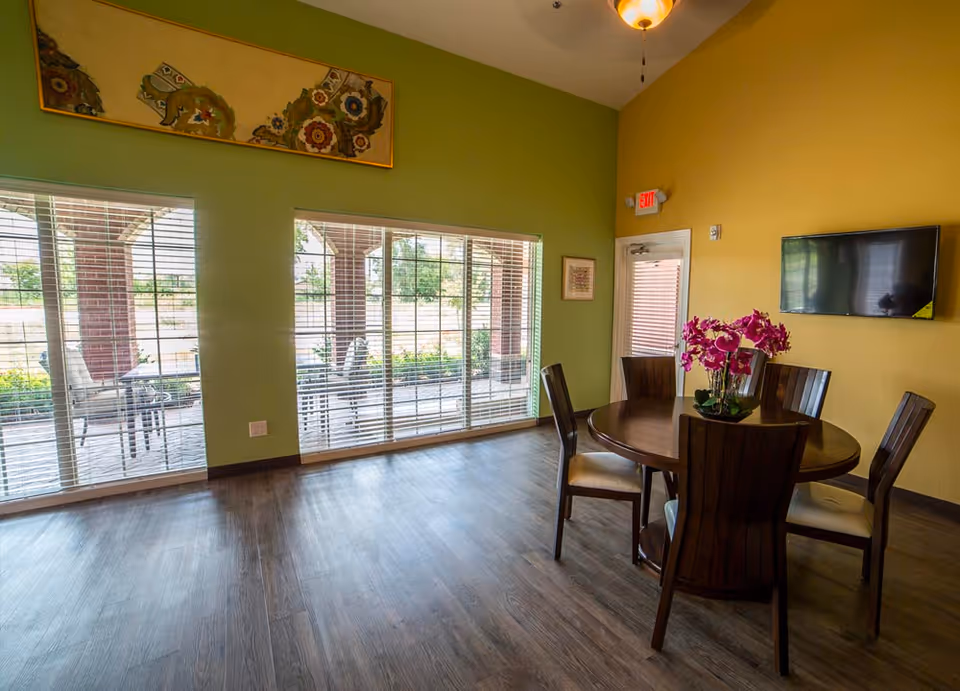 A bright room with green and yellow walls featuring a round wooden table with five chairs around it. A vase with purple flowers is placed on the table. Large windows with blinds allow natural light to enter, and outside there is a covered patio with chairs and a table. A flat-screen TV is mounted on the yellow wall, and an exit door is visible.
