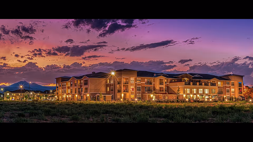 Panoramic exterior view of a multi-story senior living building lit up at sunset beneath a colorful sky.