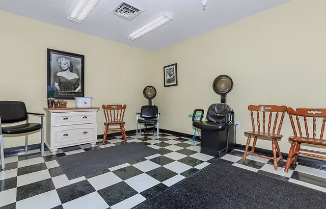 Interior of a senior living facility room with black and white checkered floor tiles, several chairs including wooden and black salon chairs, two vintage hair dryers, a white dresser with a framed Marilyn Monroe picture above it, and light yellow walls.