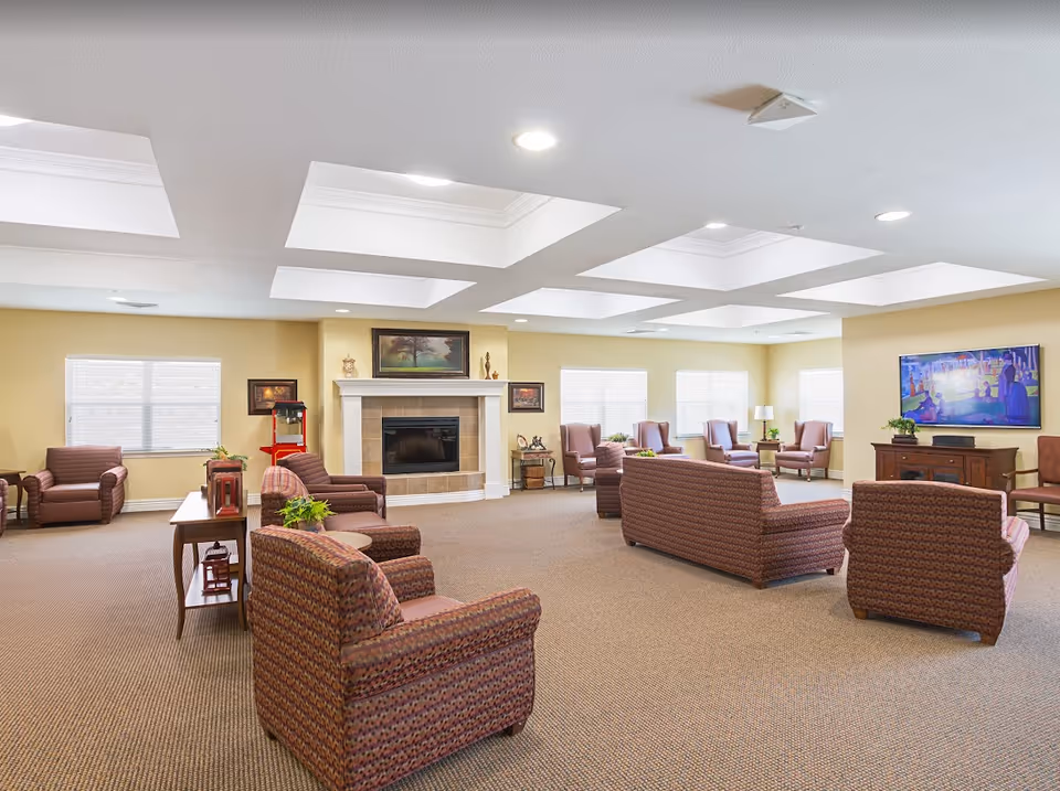A spacious and well-lit living room area in a senior living facility with multiple upholstered armchairs and sofas arranged around a central fireplace. The room has beige walls, carpeted floors, and several windows allowing natural light. There is a large flat-screen TV mounted on the wall, decorative paintings above the fireplace, and small tables with plants and lamps.