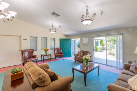 Spacious common living room with sofas and armchairs around a coffee table, chandeliers overhead and sliding glass doors to a garden.