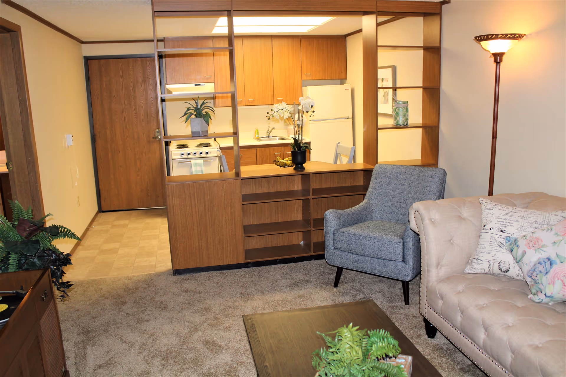 Interior view of a cozy living area in Claremont Retirement Village featuring a beige tufted sofa with floral and script-patterned pillows, a blue upholstered armchair, a wooden coffee table with a green plant, and a wooden partition with open shelving separating the living space from a small kitchen area with wooden cabinets, a white refrigerator, and a stove. A floor lamp is lit in the corner, and the entrance door is visible in the background.