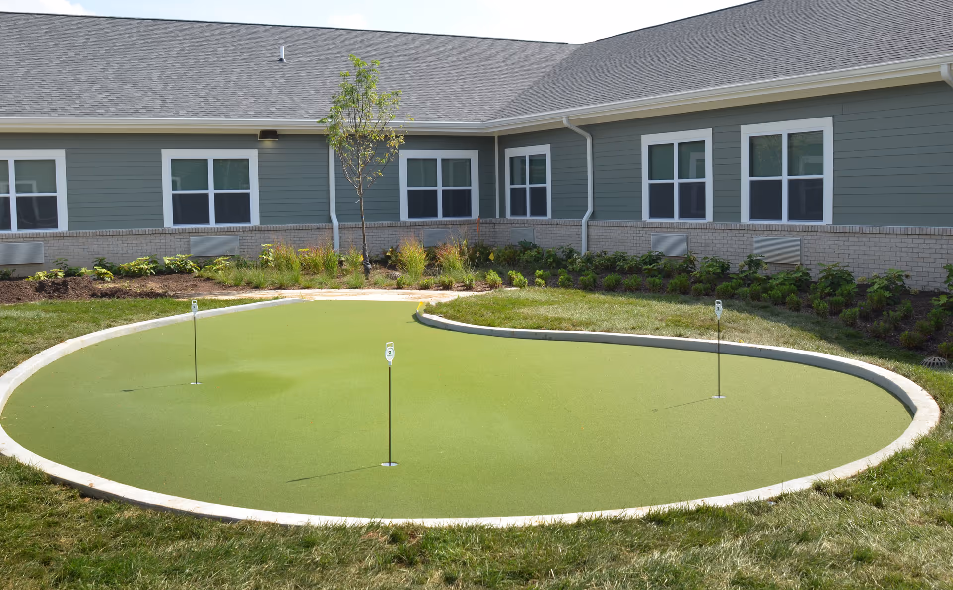 Outdoor putting green with three golf holes surrounded by grass and landscaping, adjacent to a single-story building with multiple windows and a gray roof.