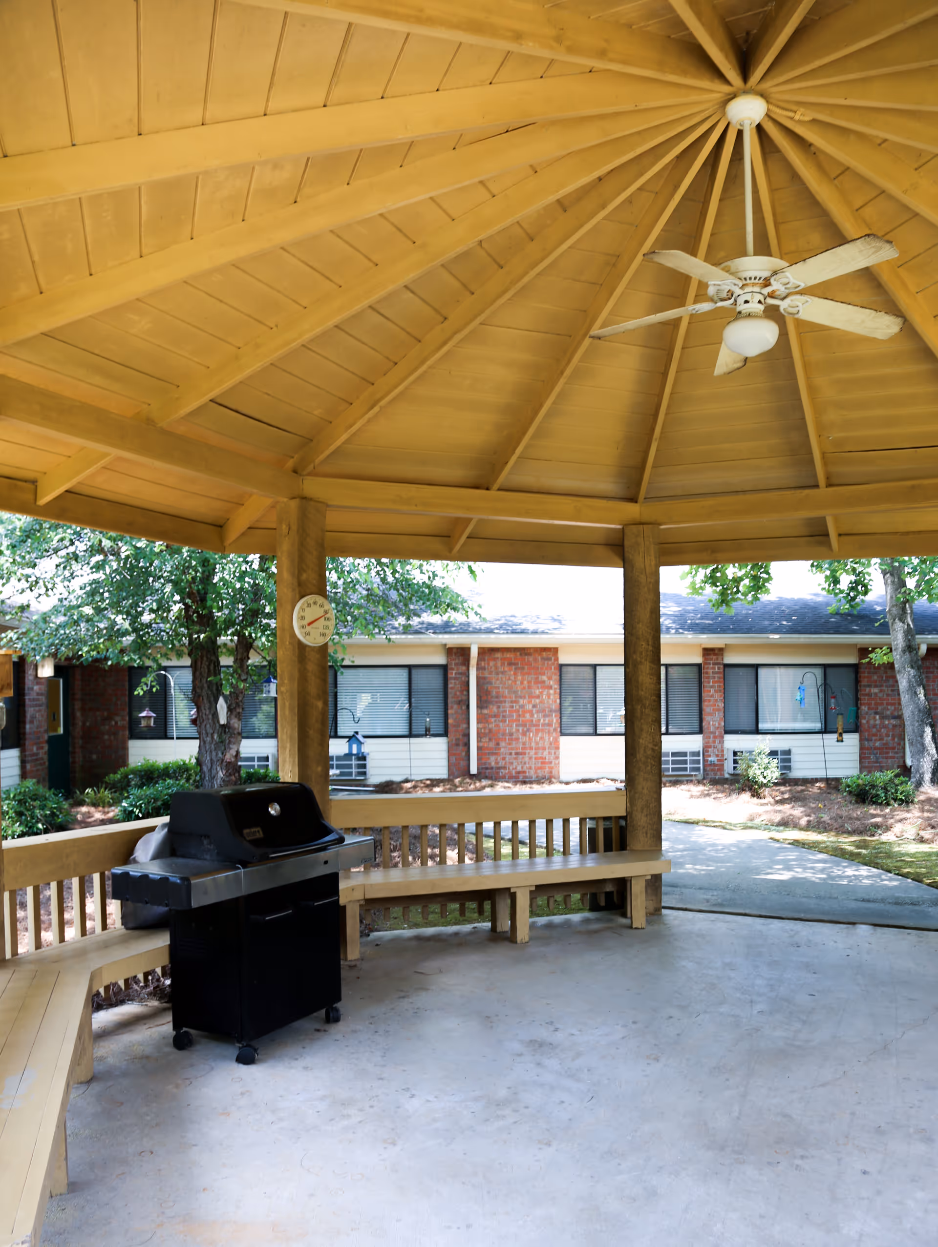 Covered outdoor gazebo area with a wooden ceiling and ceiling fan, built-in wooden benches along the perimeter, and a black grill. In the background, there is a brick building with windows and some trees and shrubs.
