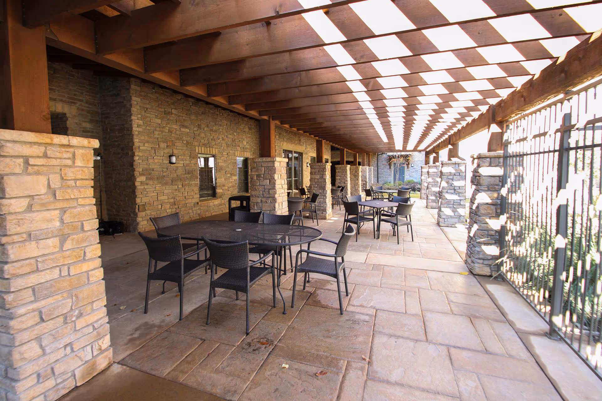 Outdoor covered patio area with stone pillars and a wooden pergola ceiling. Several black metal tables and chairs are arranged on a stone tile floor. The area is enclosed by a metal fence on one side and a stone wall with windows on the other.