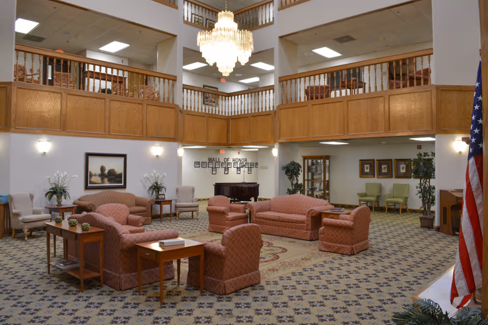 Spacious senior living facility common area with multiple upholstered sofas and armchairs arranged around wooden side tables on a patterned carpet. The room features a high ceiling with a large chandelier, wooden balcony railings on the upper floor, framed artwork on the walls, potted plants, and an American flag on the right side. A 'Wall of Honor' with framed photos is visible in the background.
