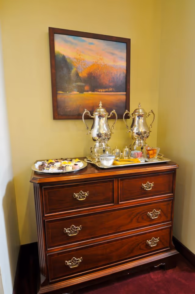 A wooden dresser with ornate handles holds two silver coffee urns, a tray with sugar packets, creamers, and a bowl of sugar, and a plate of assorted pastries. Above the dresser hangs a framed landscape painting depicting a sunset over trees and a field. The walls are painted a warm yellow color.