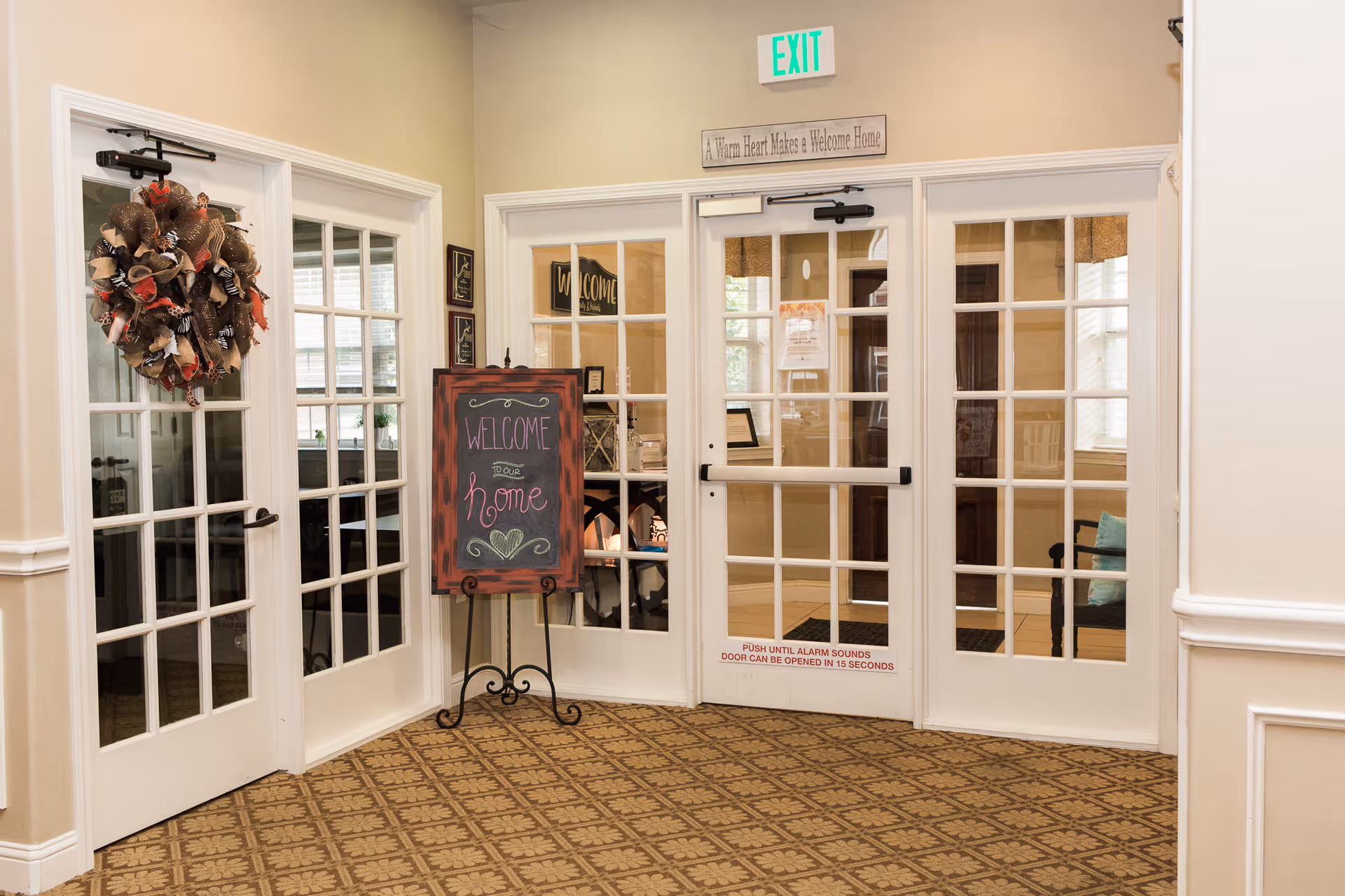 Interior view of a hallway in a senior living facility with glass-paneled double doors and a single door, a decorative wreath on the left door, a chalkboard sign that says 'Welcome to our Home', beige walls, patterned carpet, and an exit sign above the doors.