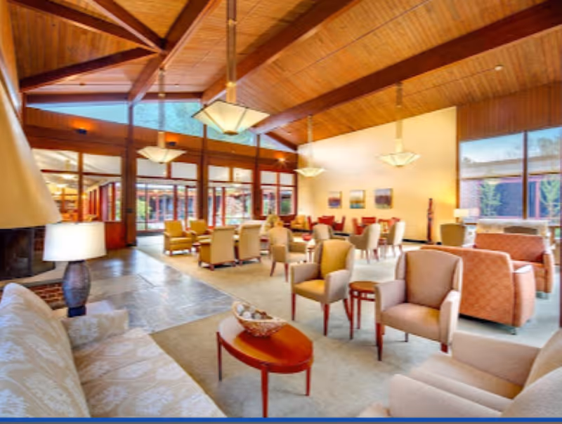 A spacious senior living facility common area with high wooden ceilings and large windows allowing natural light. The room is furnished with multiple comfortable armchairs, sofas, and small tables arranged for socializing. Pendant lights hang from the ceiling, and there are paintings on the walls. The floor is a combination of carpet and tile.