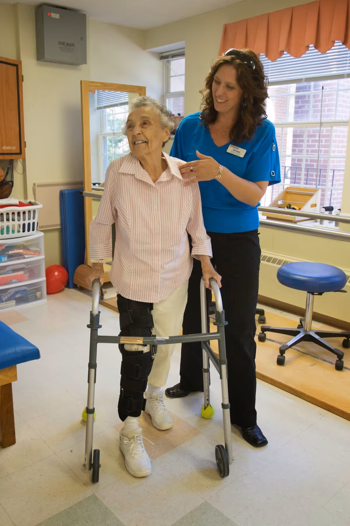 An elderly woman using a walker with a knee brace is assisted by a smiling female caregiver in a bright room with windows, exercise equipment, and a blue stool.