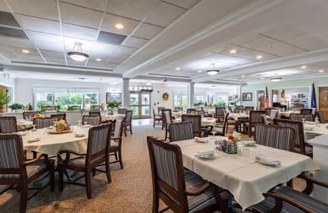 A spacious dining room in Granite Hill Estates with multiple tables covered in white tablecloths, each set with plates, bowls, and utensils. The room has large windows allowing natural light, ceiling lights, and comfortable wooden chairs with cushions. There are plants and decorative items around the room, creating a welcoming atmosphere.