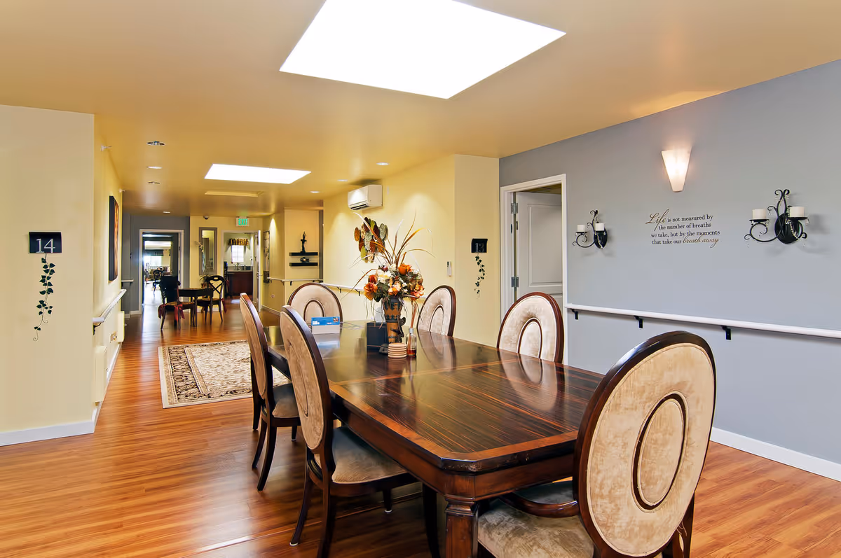 A well-lit dining area in a senior living facility with a large wooden dining table surrounded by six upholstered chairs. The room has light yellow and gray walls, wooden flooring, wall-mounted candle holders, and a decorative flower arrangement on the table. There are room number signs on the walls and a hallway leading to other rooms in the background.