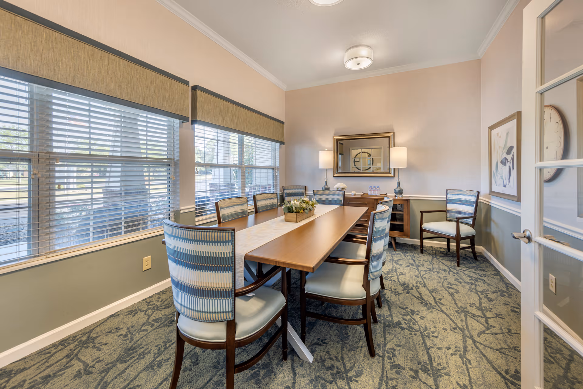 A well-lit dining room with a long wooden table surrounded by eight upholstered chairs with blue and white striped backs. The room has large windows with blinds on one side, a carpeted floor with a patterned design, a sideboard with two lamps and decorative items, a wall mirror, and framed artwork. The door to the room is partially open, showing a clock on the adjacent wall.