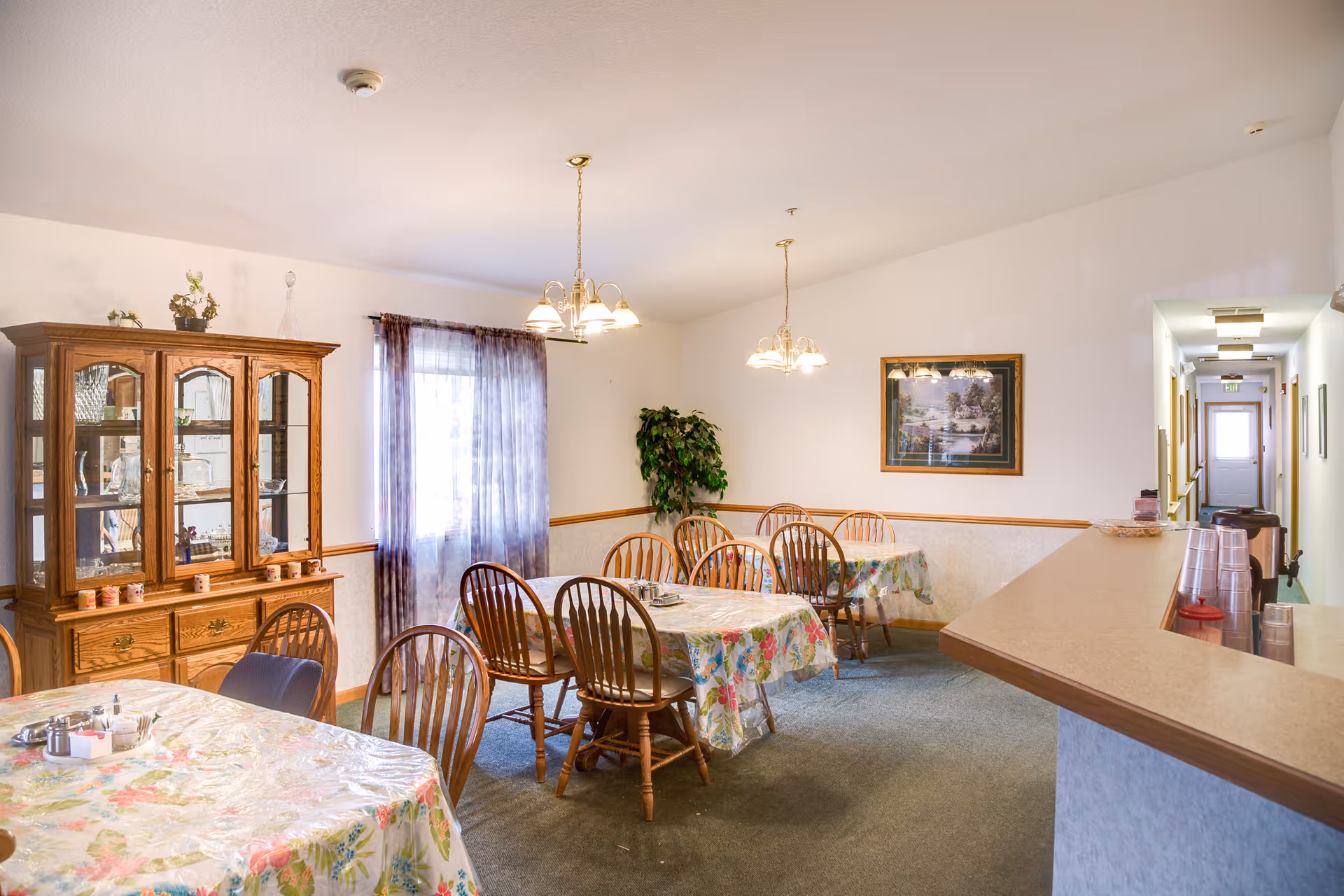 A dining room in an assisted living facility with several wooden tables covered with floral tablecloths and wooden chairs around them. There is a wooden china cabinet with glass doors on the left side, a window with sheer curtains, two hanging light fixtures, a framed picture on the wall, and a counter with plastic cups on the right side. A hallway with a door at the end is visible in the background.