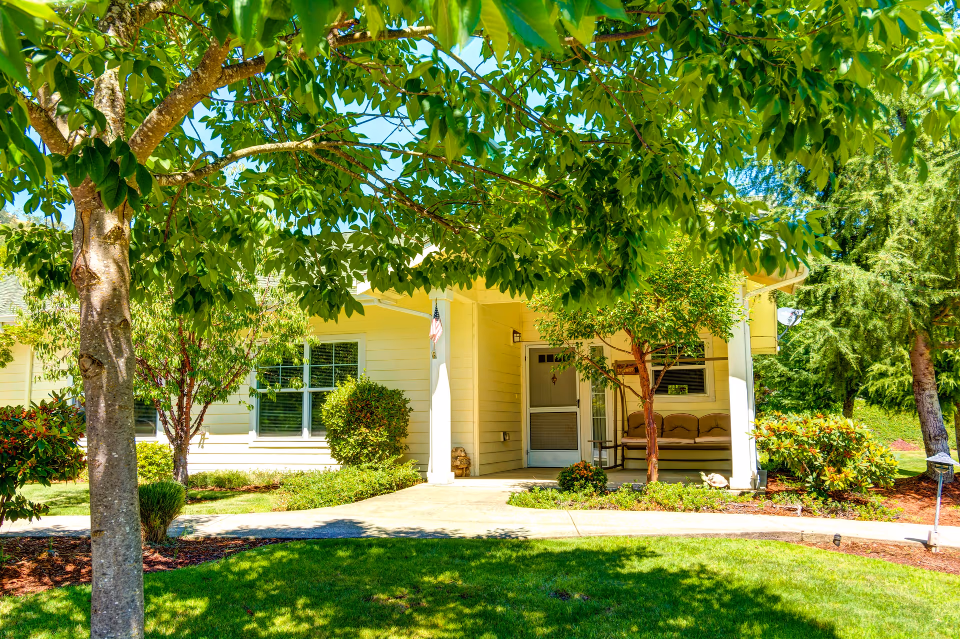 A bright and sunny outdoor view of a retirement and assisted living facility entrance with a covered porch, a bench, and well-maintained landscaping including green grass, bushes, and trees providing shade.