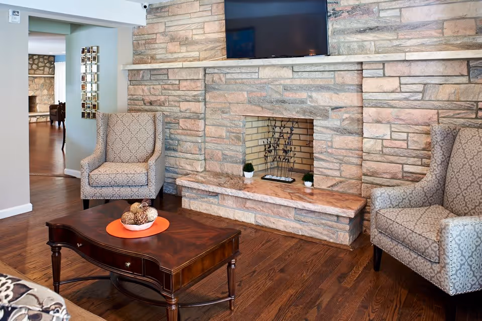 A cozy living room area with two patterned armchairs facing a wooden coffee table that has a decorative bowl with natural elements on an orange placemat. The room features a stone fireplace with a mounted flat-screen TV above it and small potted plants on the hearth. Hardwood floors and a glimpse of another room with similar stonework are visible in the background.