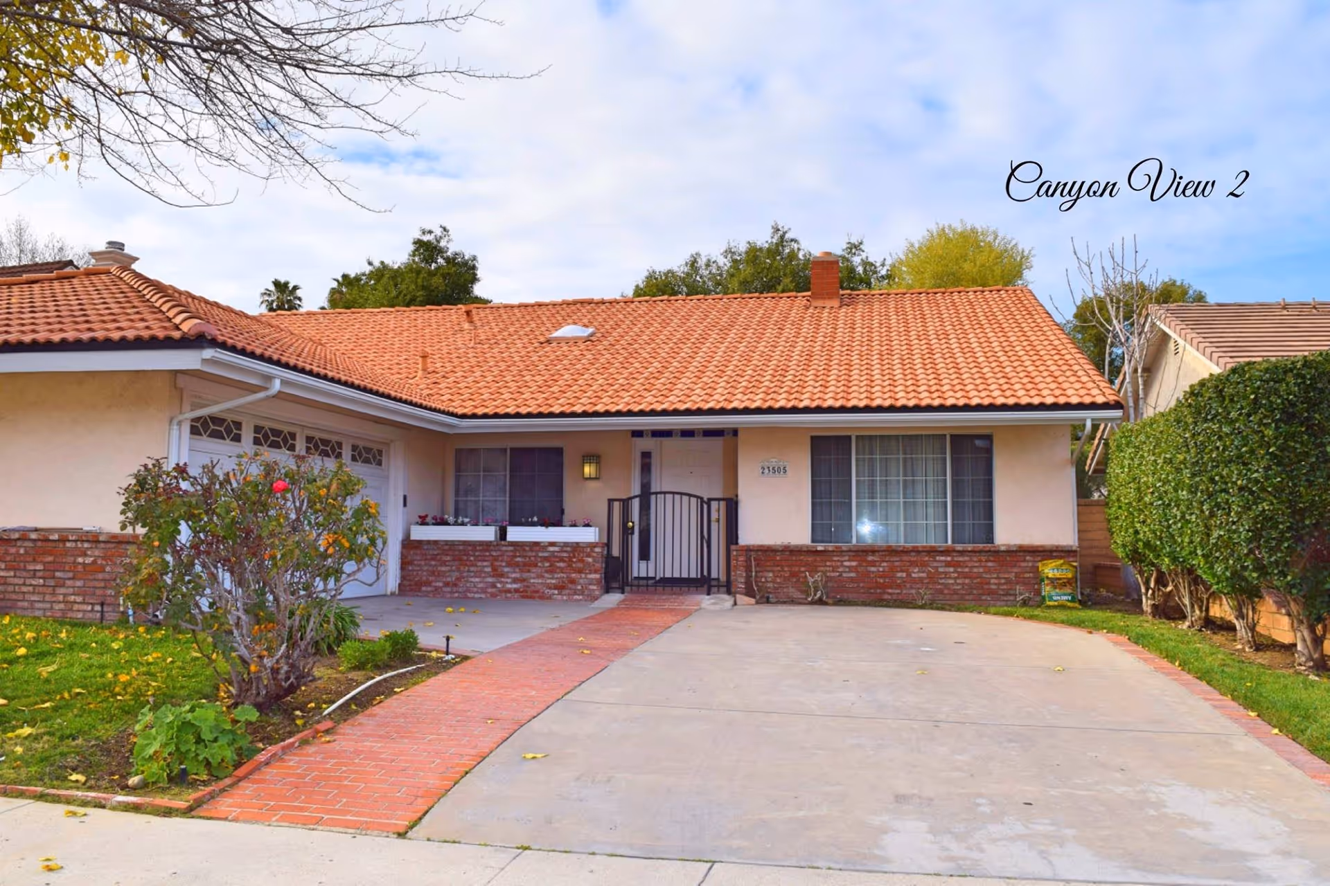 Front exterior view of a single-story residential building with a red tiled roof, beige walls, a brick lower facade, a driveway, and a small garden with bushes and trees. The sky is partly cloudy.
